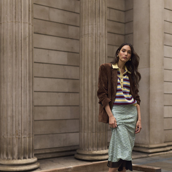 A woman stands on a wooden bench in front of large stone columns, wearing a brown suede jacket over a striped knit top, a light mint midi skirt, yellow socks, and brown suede sneakers.