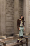 A woman stands on a wooden bench in front of large stone columns, wearing a brown suede jacket over a striped knit top, a light mint midi skirt, yellow socks, and brown suede sneakers.