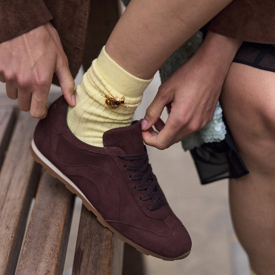 A close-up lifestyle shot of a person sitting on a wooden bench, tying the laces of a dark brown suede sneaker with a pale yellow sock featuring a small bee brooch.