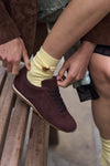 A close-up lifestyle shot of a person sitting on a wooden bench, tying the laces of a dark brown suede sneaker with a pale yellow sock featuring a small bee brooch.