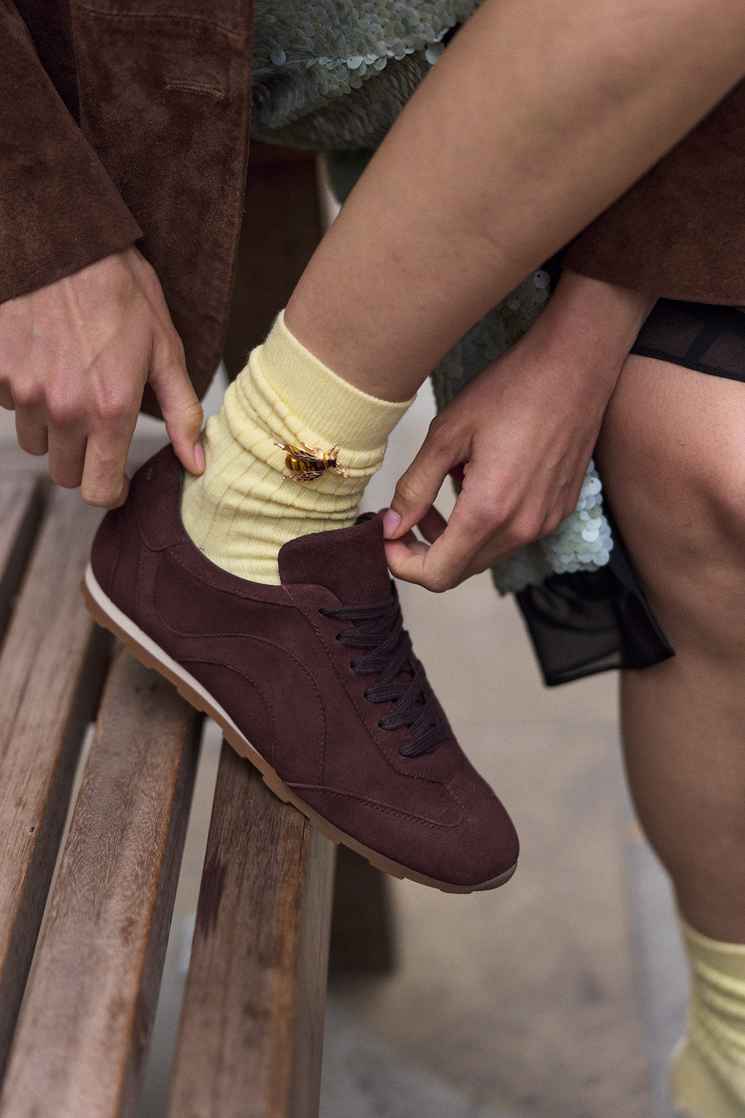 A close-up lifestyle shot of a person sitting on a wooden bench, tying the laces of a dark brown suede sneaker with a pale yellow sock featuring a small bee brooch.