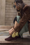 A woman sitting on a wooden bench wearing a brown suede jacket, a mint patterned skirt, and dark chocolate brown sneakers with yellow socks.