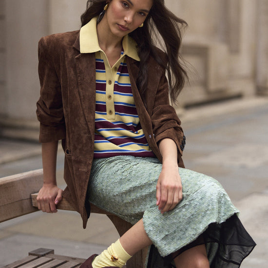 A woman models a brown suede jacket over a striped polo shirt, a textured mint green skirt, and brown suede trainers while sitting on a wooden bench in an outdoor urban setting.