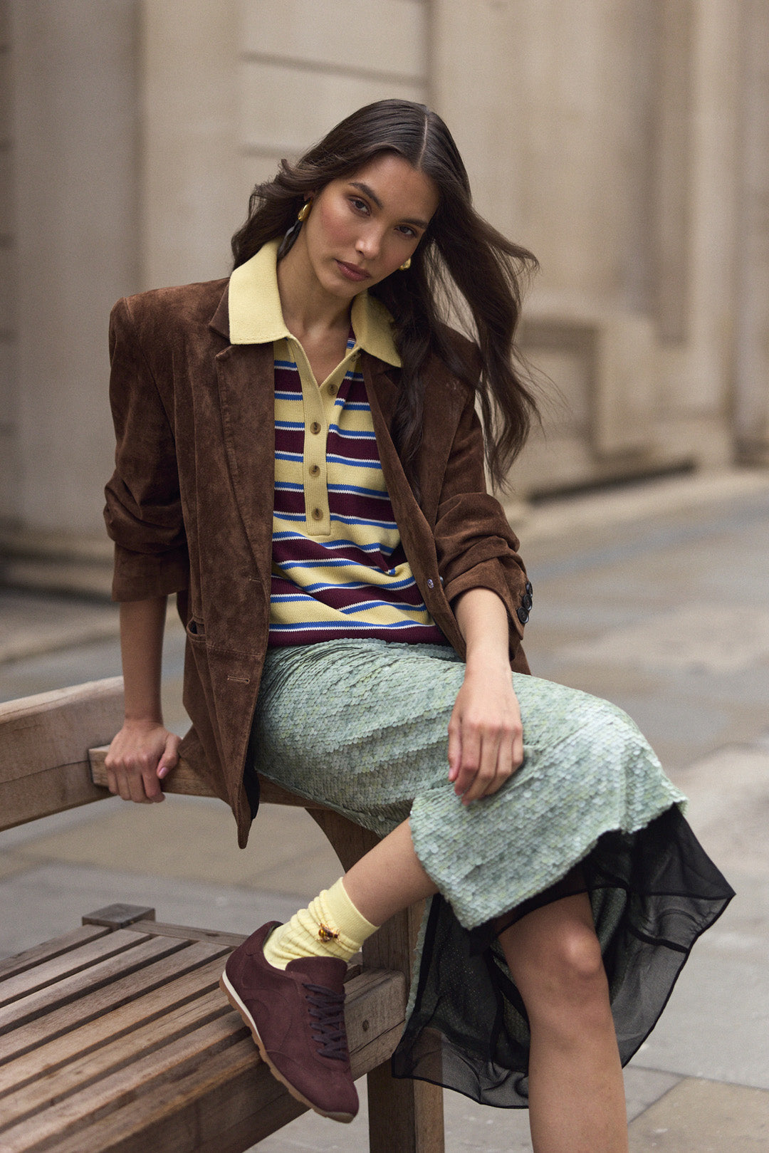 A woman models a brown suede jacket over a striped polo shirt, a textured mint green skirt, and brown suede trainers while sitting on a wooden bench in an outdoor urban setting.