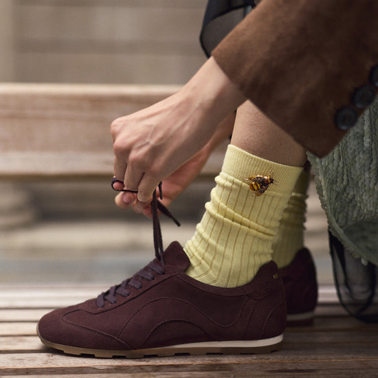 A person sitting on wooden steps ties the laces of a dark brown suede sneaker while wearing a pale yellow ribbed sock with a small gold bee brooch detail.