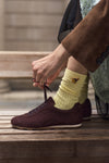 A person sitting on wooden steps ties the laces of a dark brown suede sneaker while wearing a pale yellow ribbed sock with a small gold bee brooch detail.