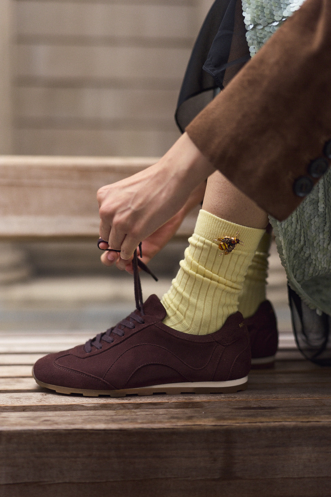 A person sitting on wooden steps ties the laces of a dark brown suede sneaker while wearing a pale yellow ribbed sock with a small gold bee brooch detail.