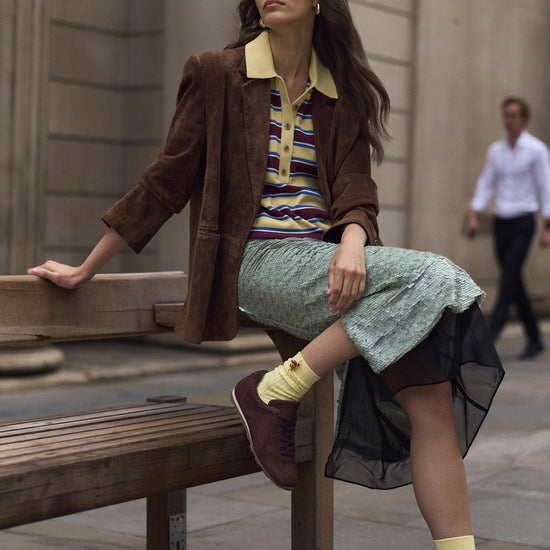 A woman sits on a wooden bench in an outdoor urban setting, wearing a brown suede jacket, a striped knit polo shirt, a light green patterned skirt, and chocolate brown suede sneakers with yellow socks.