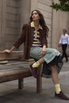A woman sits on a wooden bench in an outdoor urban setting, wearing a brown suede jacket, a striped knit polo shirt, a light green patterned skirt, and chocolate brown suede sneakers with yellow socks.