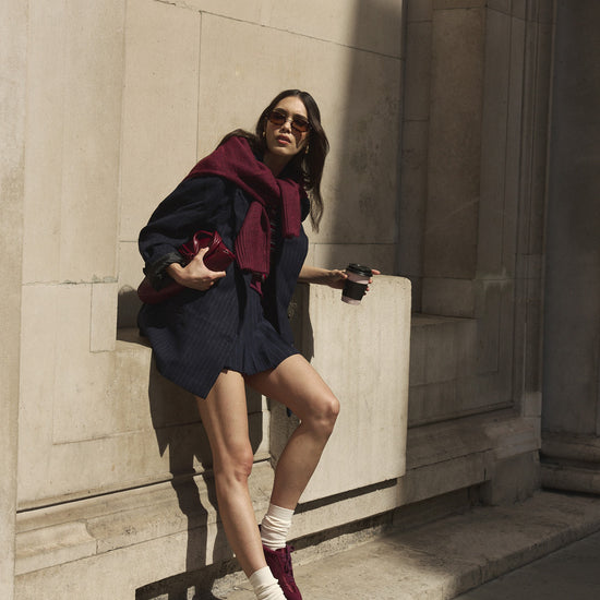 A woman posing outdoors against a stone wall, wearing a dark navy outfit, a deep burgundy jumper round her shoulders, cream socks, and burgundy satin and suede trainers.