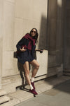 A woman posing outdoors against a stone wall, wearing a dark navy outfit, a deep burgundy jumper round her shoulders, cream socks, and burgundy satin and suede trainers.