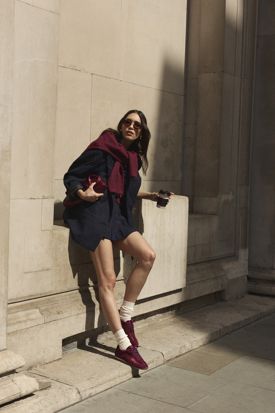 A woman posing outdoors against a stone wall, wearing a dark navy outfit, a deep burgundy jumper round her shoulders, cream socks, and burgundy satin and suede trainers.
