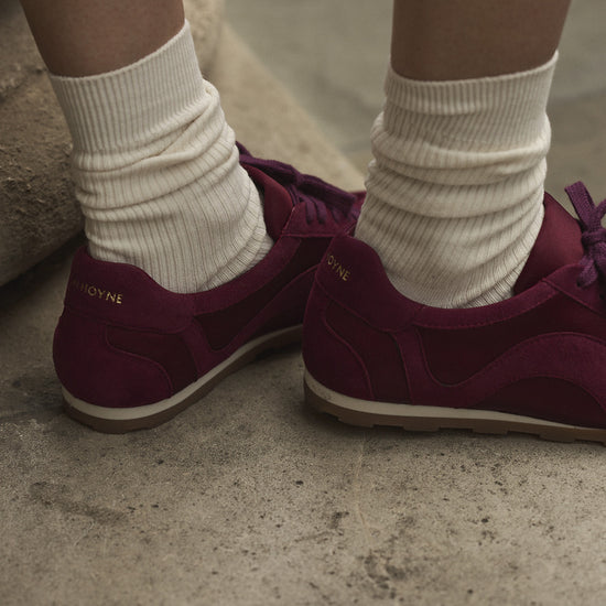 A close-up of the the back of a person's burgundy satin and suede sneakers, paired with cream crew socks, standing on stone steps.