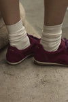A close-up of the the back of a person's burgundy satin and suede sneakers, paired with cream crew socks, standing on stone steps.