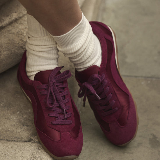 A close-up of a person's feet wearing burgundy satin and suede sneakers with matching laces, paired with cream crew socks, standing on stone steps.