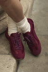 A close-up of a person's feet wearing burgundy satin and suede sneakers with matching laces, paired with cream crew socks, standing on stone steps.
