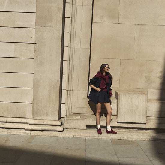 A woman wearing a dark navy mini skirt, navy blazer, and burgundy satin trainers stands against a large beige stone building wall in winter sunlight.