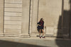 A woman wearing a dark navy mini skirt, navy blazer, and burgundy satin trainers stands against a large beige stone building wall in winter sunlight.