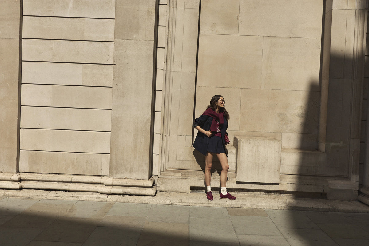 A woman wearing a dark navy mini skirt, navy blazer, and burgundy satin trainers stands against a large beige stone building wall in winter sunlight.