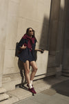 A woman posing outdoors against a stone wall, wearing a dark navy pleated mini skirt, a dark navy blazer, a deep burgundy jumper over her shoulders, white socks, and burgundy sneakers.